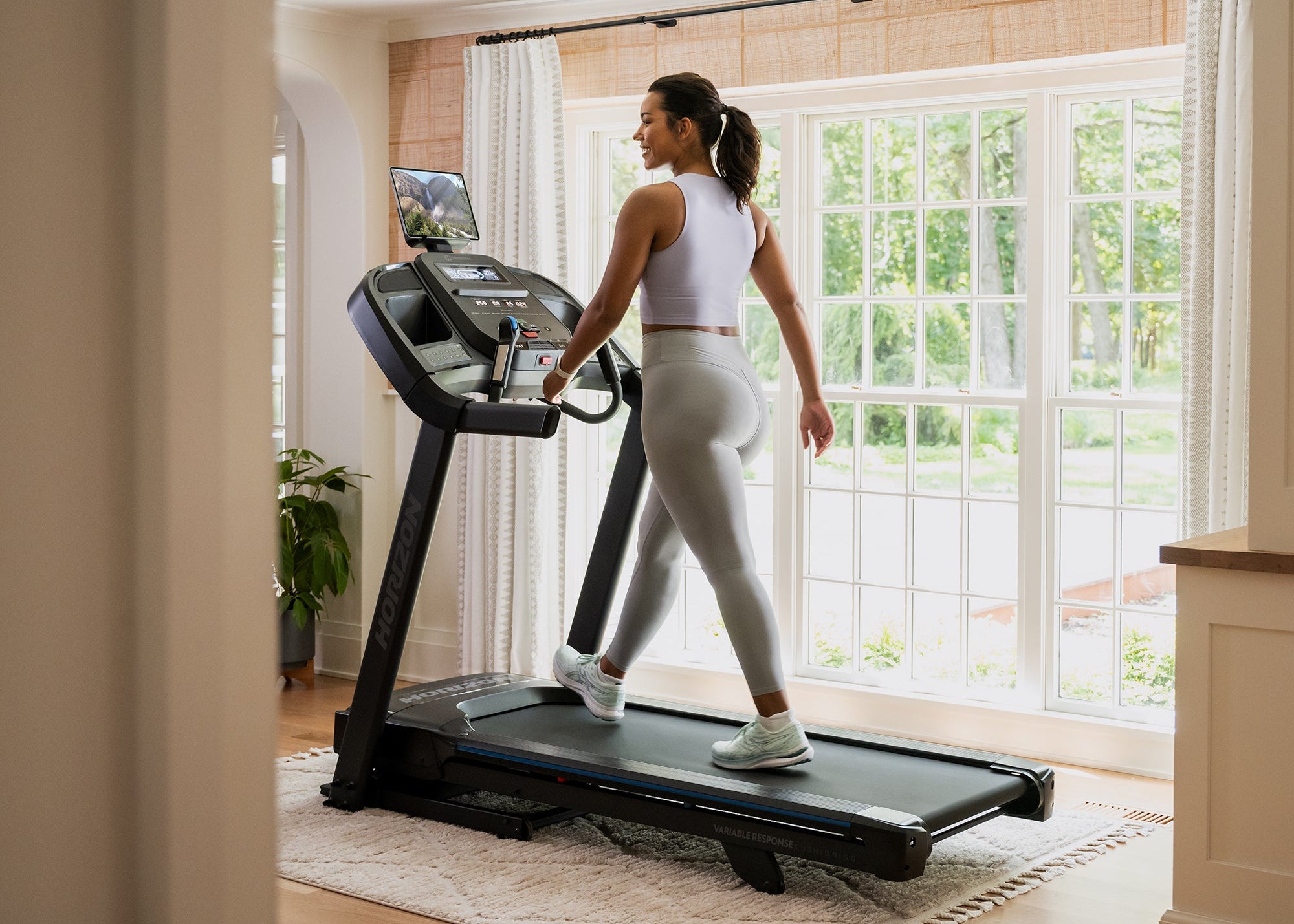 Woman using a treadmill in a home setting with large windows.