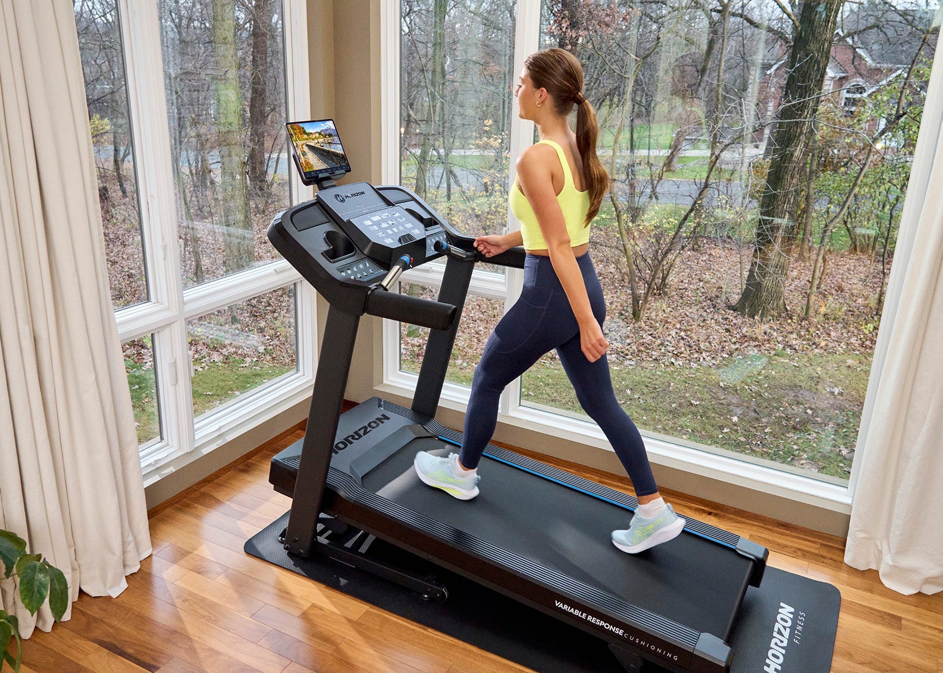 Woman using a treadmill in a home setting with large windows.